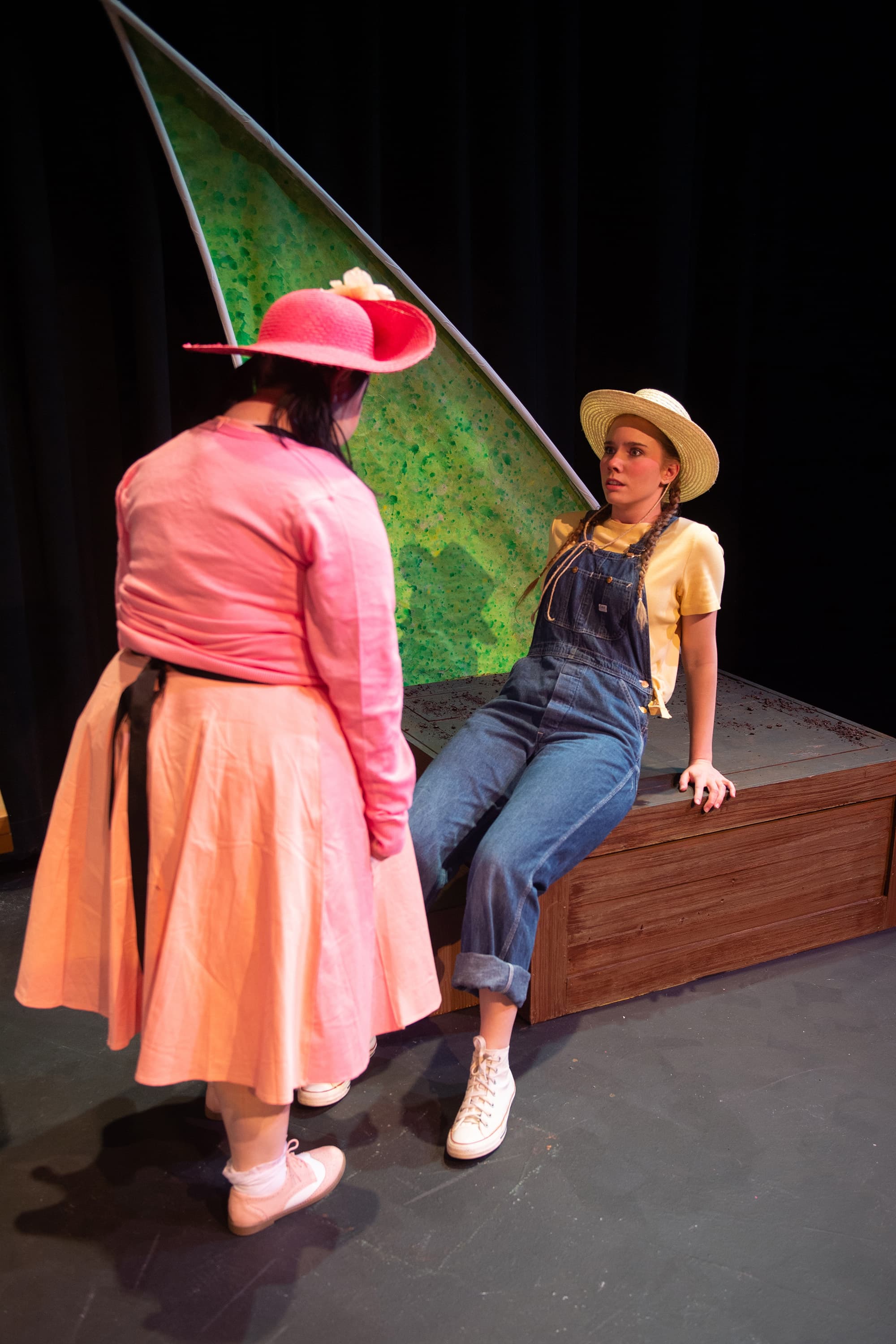 Two female students on stage during a theatre performance. One is sitting and wearing overalls. The other is standing and looking at the other, wearing all pink.